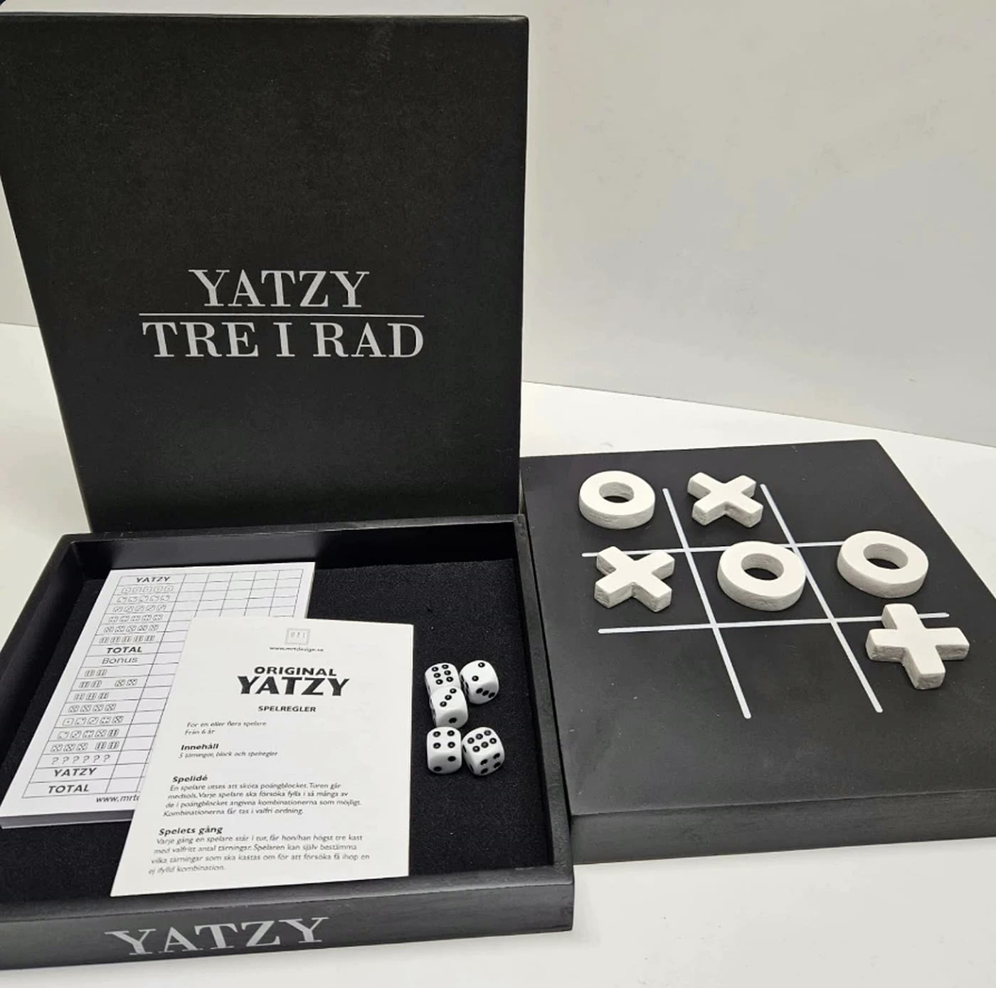 Öppen Yatzy Tre i rad-box med scoreblock, spelregler och tärningar, bredvid locket med vita pjäser på brädet